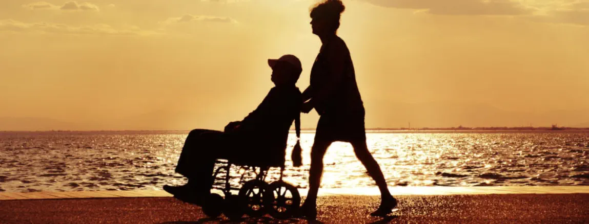 Eine Frau schiebt ein Mann im Rollstuhl am Meer in den Sonnenuntergang